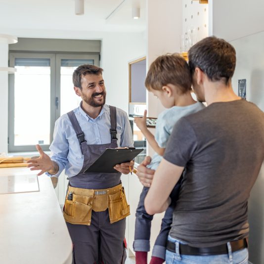 Professional plumber in uniform talking to male client and his small son indoors.